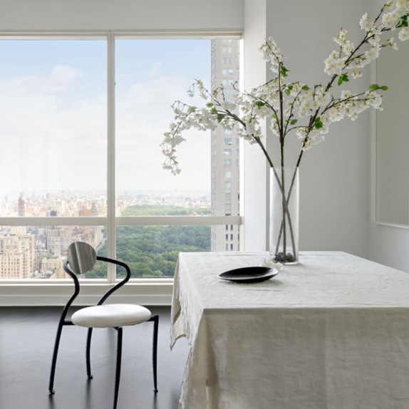 Minimalist, contemporary penthouse dining room interior by Orlie Katsiris Staging & Interiors featuring a linen-draped dining table, sculptural black chair, oversized glass vase with flowering branches, and expansive city skyline views.
