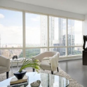 Contemporary, neutral penthouse living room interior by Orlie Katsiris Staging & Interiors featuring sculptural accent chairs, a round glass coffee table, organic decor, and floor-to-ceiling windows overlooking the Manhattan skyline.